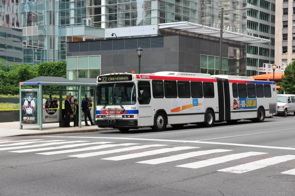 A bus stop in Philadelphia, where people wait under a glass shelter for a bus as it arrives.